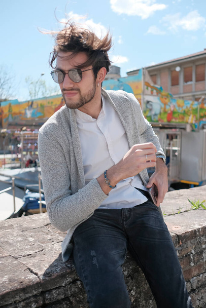 handsome Italian dark-haired guy with jacket sitting on low wall in sunny day. High quality photo - Foto, Imagen