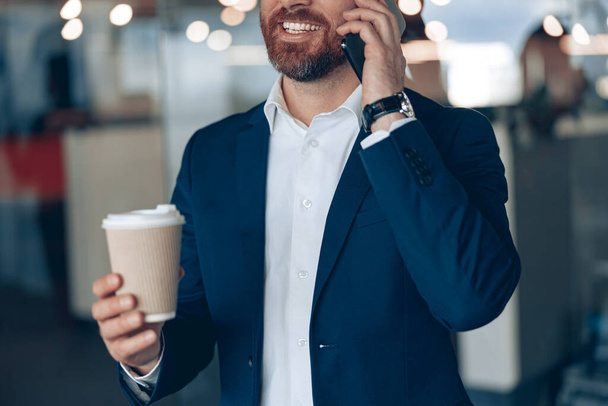 Primo piano di un uomo d'affari sorridente con tazza di caffè da asporto che parla al cellulare con un socio in affari - Foto, immagini