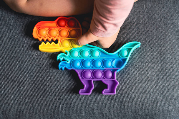 Toddler girl with popit toy in rainbow colours. Dino shape silicone toy for stress relief. Bubbles sensory trendy fidget. Child playing at home in candid style - 写真・画像