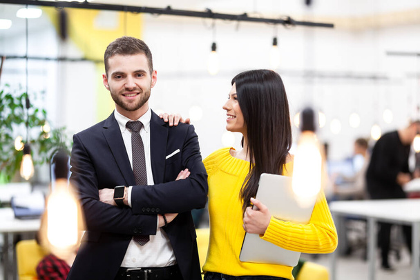 Beautiful Caucasian dark haired girl in yellow posing with businessman wearing suit. Happy colleagues with a laptop in a modern office - Fotó, kép