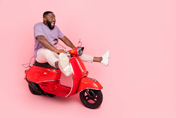 Photo of pretty impressed guy wear purple t-shirt riding moped empty space isolated pink color background - Фото, изображение