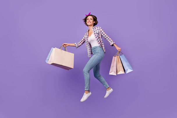 Photo of hurrying sweet lady dressed checkered shirt jumping high holding bargains isolated purple color background - Foto, Imagen