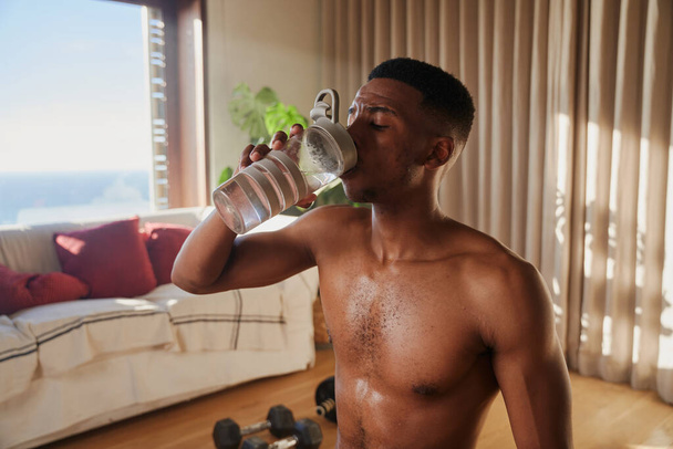Young African American Black male drinking water after workout at home in living room. Keeping fit, hydrated and healthy. - Fotografie, Obrázek