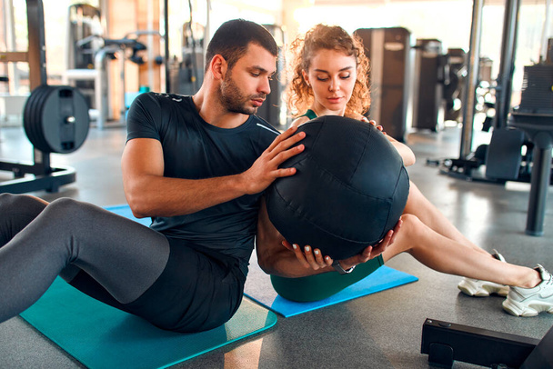 Slender caucasian woman and muscular man in sportswear doing exercises with a heavy medicine ball on a rubber mat in a gym club. The concept of sports and recreation. - 写真・画像
