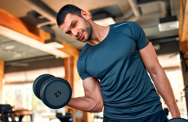 Hombre atlético musculoso en ropa deportiva haciendo un ejercicio con pesas para bíceps en un club de gimnasia. El concepto de deporte y recreación. - Foto, Imagen