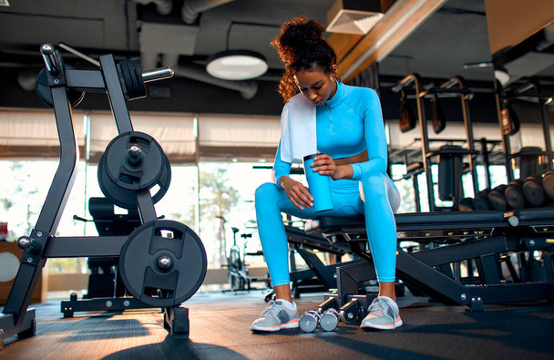Slim athletic african american woman in sportswear is tired drinking water or protein shake while relaxing after workout in gym club. The concept of sports and recreation. - Photo, Image