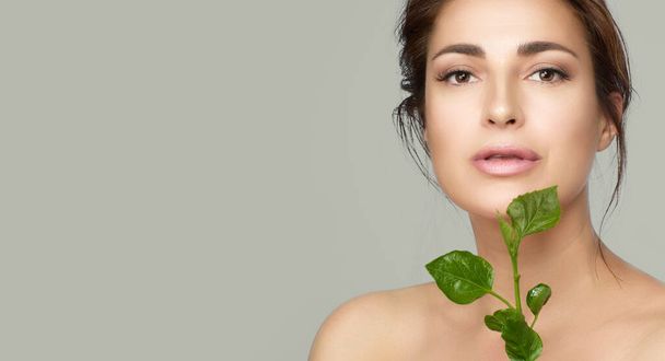 Natural beauty with green leaves. Beautiful woman with flawless skin and nude makeup on her hydrated and glowing skin. Natural cosmetics and skincare concept. Beauty portrait over greenish grey background - 写真・画像