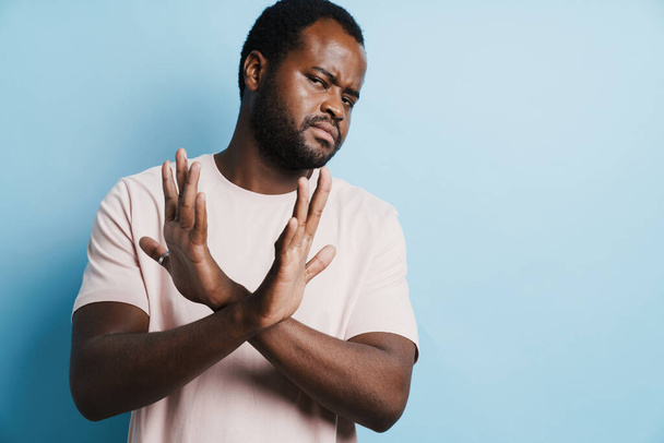 Black bristle man in t-shirt frowning and showing stop gesture isolated over blue background - Fotó, kép