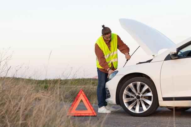 Молодий чоловік на жилеті з червоним попереджувальним трикутником знак від дороги намагається полагодити зламану машину
 - Фото, зображення