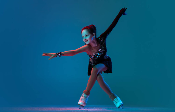 Dynamic portrait of young girl, female figure skater in black stage dress skating isolated on blue background in neon light. Concept of sport, beauty, active lifestyle. - Photo, image