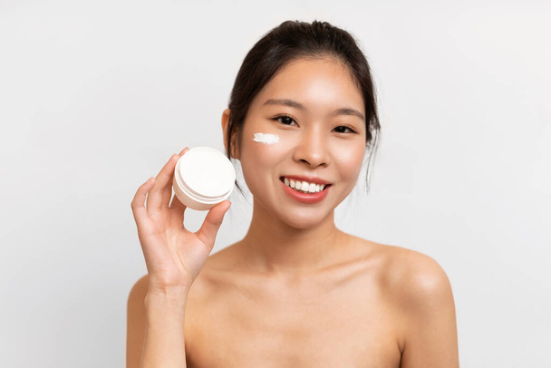 Portrait of Asian woman with cream on cheek holding jar - Фото, зображення