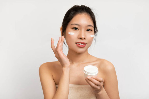Portrait of Asian lady with cream on cheek holding jar - Fotó, kép