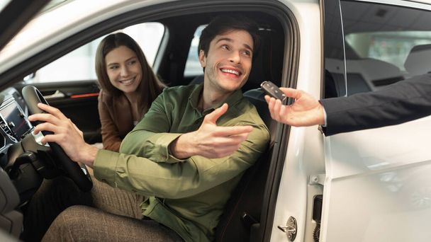 Feliz pareja millennial tomando la llave del coche del vendedor de automóviles, sentado dentro del automóvil moderno en la concesionaria, panorama - Foto, Imagen