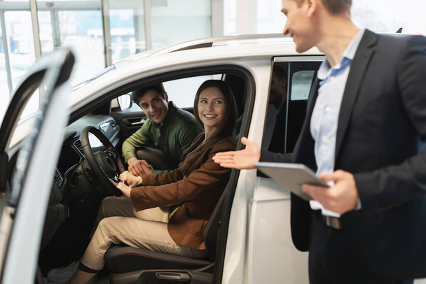 Millennial car salesman discussing purchase of new auto with happy young couple after test drive at dealership centre - Foto, Imagem