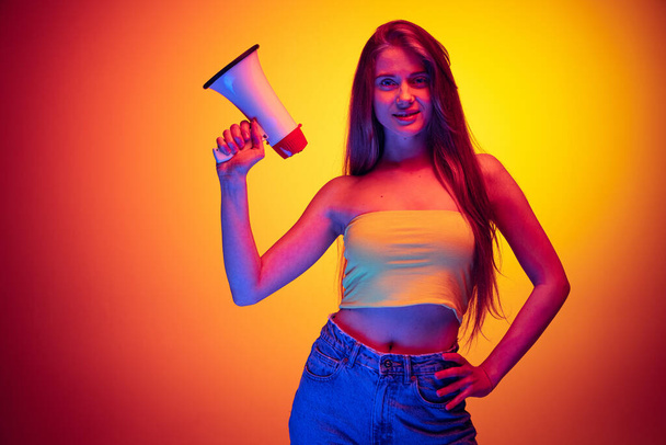 Portrait of young beautiful girl posing with megaphone isolated over gradient red yellow background in neon light - Foto, Bild