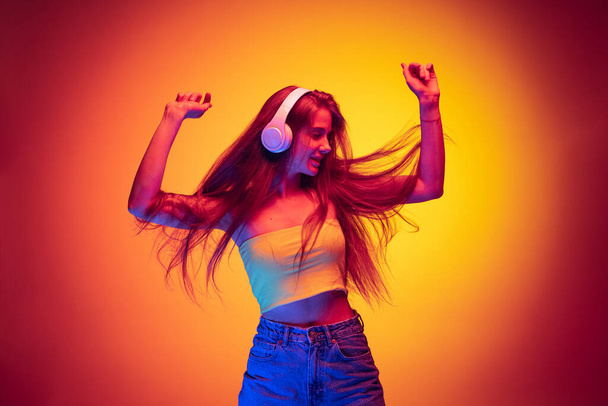 Dynamic portrait of young beautiful girl listening to music in headphones and dancing isolated over gradient red yellow background in neon - Foto, Bild