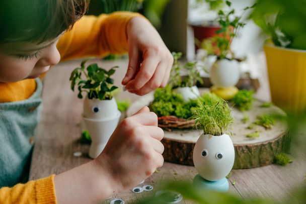 Little girl decorating eggshells with grass with toy stickers eyes on the wooden table at home. Creative fun DIY idea for festive Easter decoration. Selective focus. - Photo, Image