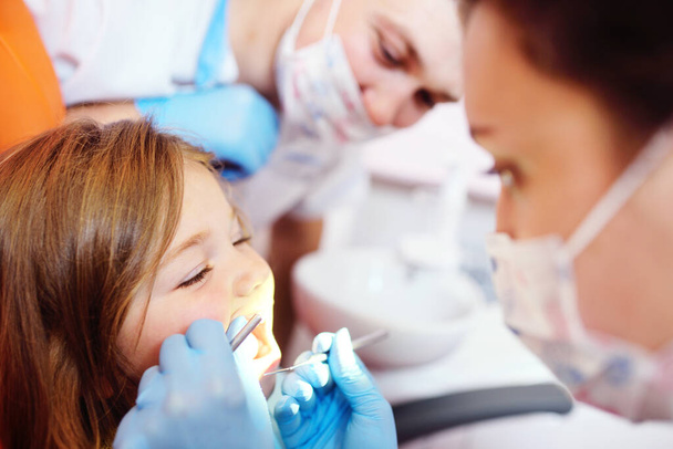 young dentists, a man and a woman, examine the teeth of a childs patient - a little pretty girl who is sitting in an orange dental chair. - Фото, зображення