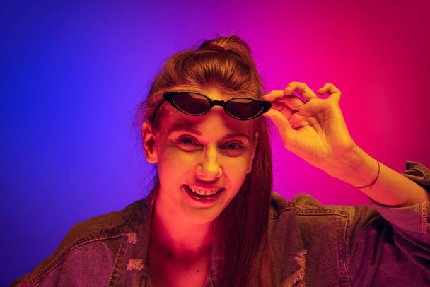Porrtrait of young smiling girl in denim jacket laughing at camera, posing isolated over gradient pink blue background in neon light - Fotó, kép