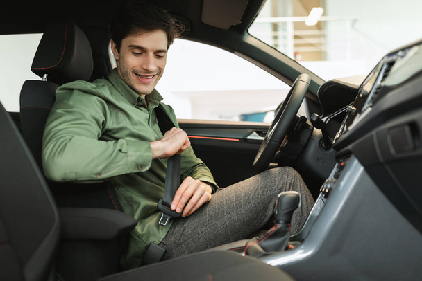 若い男性のクライアントは、ブランドの新しい車の運転席に座って、シートベルトを締結し、自動車販売店でテストドライブに行く - 写真・画像