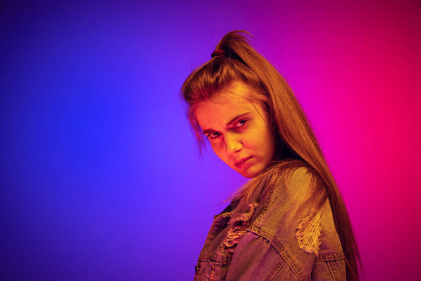 Portrait of young girl in denim jacket making serious facial expression, looking at camera isolated over blue pink background in neon - Фото, изображение