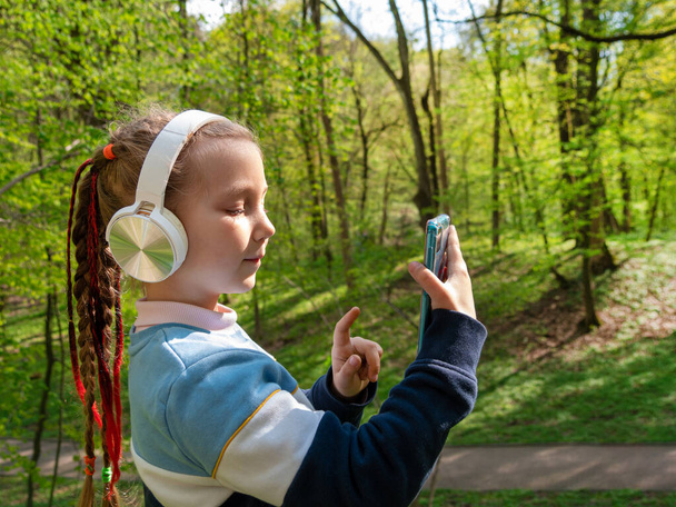 Adorable child girl in headphones learning language listening to music using smartphone outdoor sunny day. Blogger kid with gadget distance school online lesson chat cell phone conference in the park. - 写真・画像