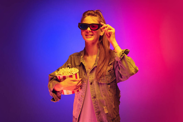 Portrait of young girl in denim jacket wearing 3D cinema glasses and holding popcorn basket isolated over gradient pink purple background in neon - 写真・画像