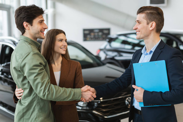 Giovane coppia caucasica stringendo la mano con il venditore di auto, l'acquisto di auto nuove in concessionaria - Foto, immagini