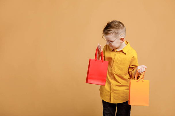 Shopping on black friday. Little boy holding shopping bags on yellow background. Shopper with many colored paper bags. Holidays sales and discounts. Cyber monday. High quality photo. - 写真・画像