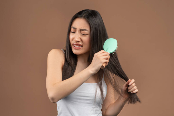 Cosmetics for problems, dry and brittle hair. Young korean lady cannot comb her long hair, brown studio background - Foto, Imagem