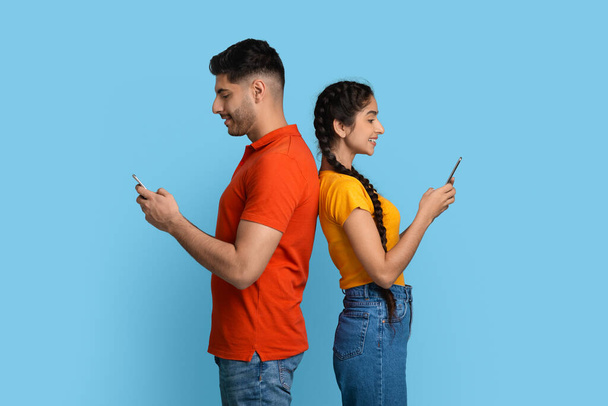 Fröhliches Paar aus dem Nahen Osten, das Rücken an Rücken steht und Smartphones benutzt - Foto, Bild