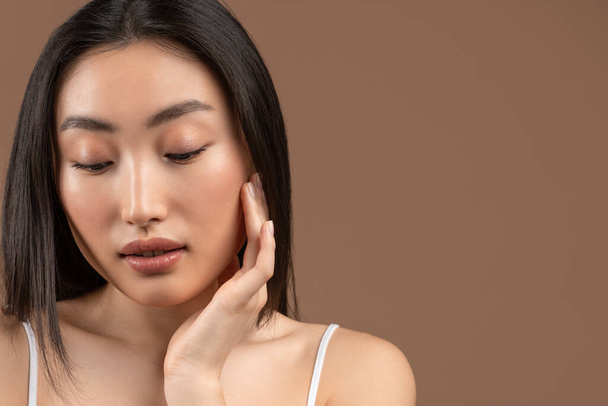 Natural skincare. Young pretty asian woman caressing her smooth soft skin, brown background, copy space - Foto, imagen