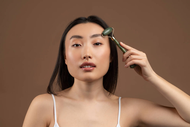 Beautiful asian lady using jade face roller on her flawless skin, looking at camera while posing over brown background - Zdjęcie, obraz