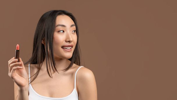 Aantrekkelijke Aziatische vrouw houdt lippenstift in de hand, kijkt opzij naar de vrije ruimte en glimlacht over bruine achtergrond, panorama - Foto, afbeelding