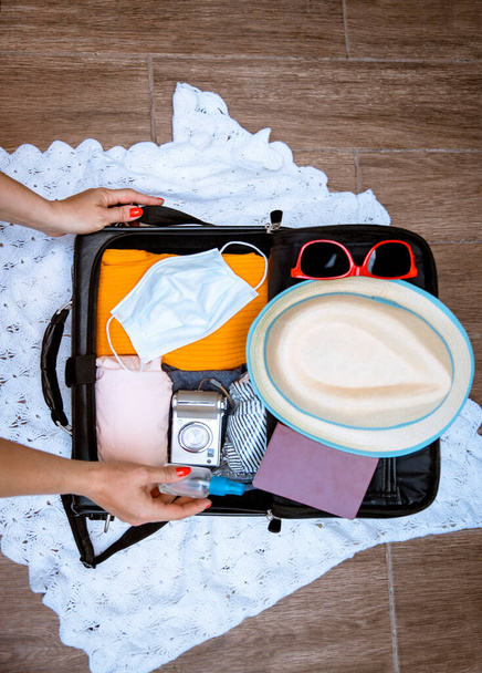 Packing suitcase luggage with face mask and sanitizer with clothes for new normal travelling during Coronavirus pandemic. Cut out image of female hands preparing for the vacation travel. Top view flat - Photo, image