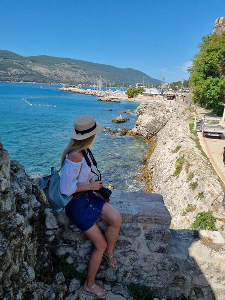 young tourist girl with hat and sunglasses on the beach makes photos of the nature and enjoying and looking at the view on the sea, in Herceg Novi, Montenegro - Фото, изображение