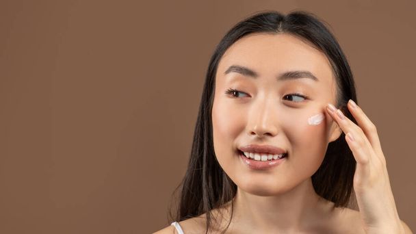 Female skincare concept. Happy asian woman applying moisturizer cream, brown background, copy space - Photo, Image