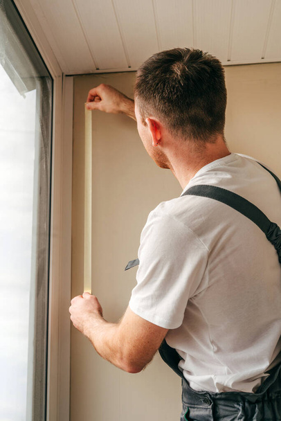 Jongeman die een overall draagt en plakband aanbrengt voordat hij een raam afsluit met waterdichte siliconen ketel op het balkon. Selectieve focus. - Foto, afbeelding