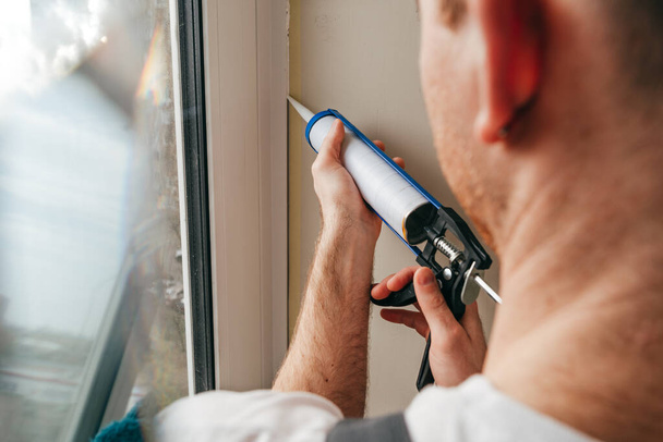 Giovane uomo che indossa tuta sigillare crepe tra finestra e finiture utilizzando calafatello in silicone impermeabile sul balcone. - Foto, immagini