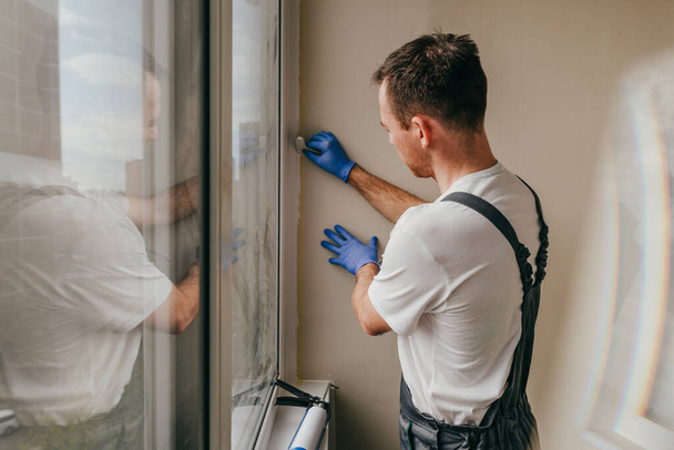 Young man wearing overalls sealing cracks using waterproof silicone caulk on the balcony. Removing the sealant using a rubber spatula. Selective focus. - Foto, Imagem