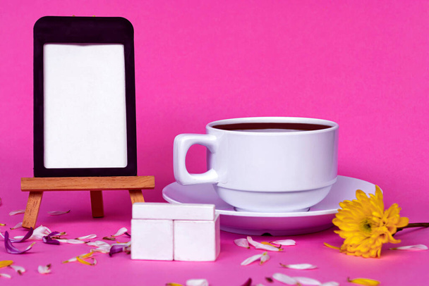 Chalkboard with white coffee cup, wooden block and flowers on pink background. - Фото, изображение