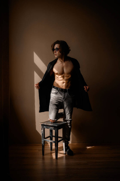A handsome guy in a black long cardigan and torn jeans stands near a stool - Foto, Imagem