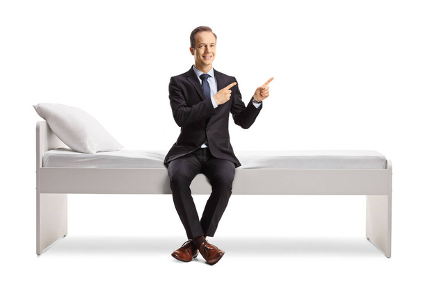 Businessman sitting on a bed and pointing with both fingers to the side isolated on white background - Photo, image