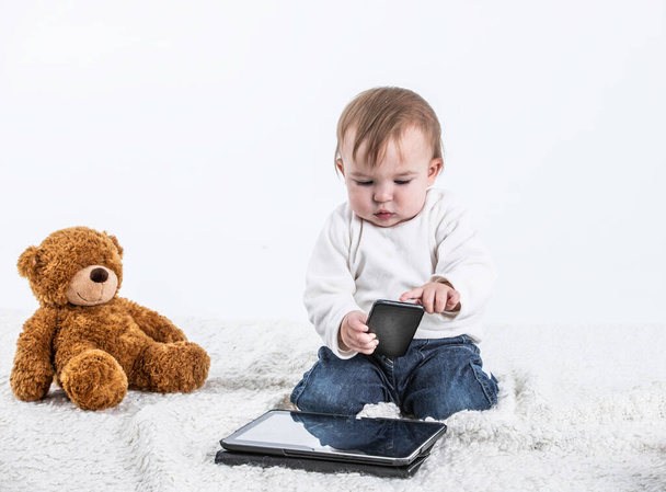 Foto de estudio en stock con un fondo blanco de un bebé tocando la pantalla de una tableta con un oso de peluche en el lateral y un recipiente con asas para beber
. - Foto, Imagen