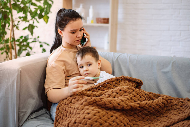 A sad sick boy is lying on the couch, feels a fever, has a cold, flu. The worried mother presses the phone to her ear and calls the family therapist after measuring the childs temperature - Photo, Image
