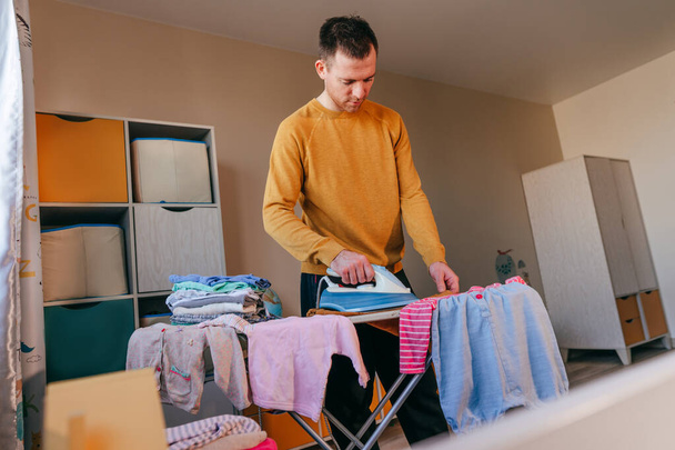 Young man ironing baby clothes in the children's room. Father doing chores at home. Domestic life concept. Selective focus. - Foto, afbeelding