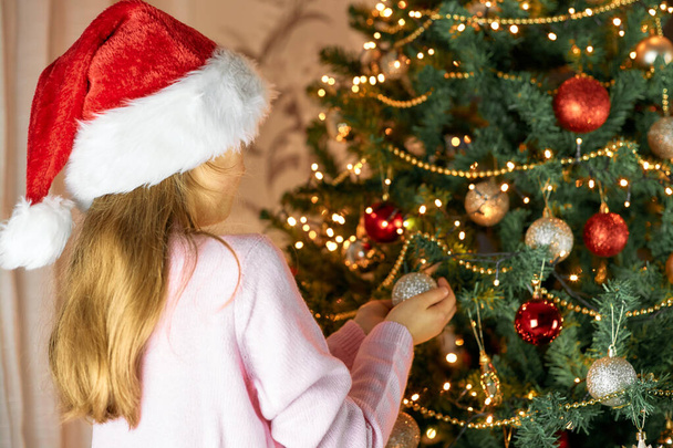 Tyttö punaisessa Santa hattu koristaa joulukuusi pallot ja seppeleitä. Valmistaudun uuteen vuoteen ja jouluun. Odotan ihmettä. - Valokuva, kuva