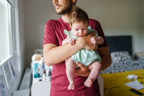 Caucasian man holding his baby girl child daughter by the window at home in room in summer day - parenthood father's day concept real people front view - Valokuva, kuva