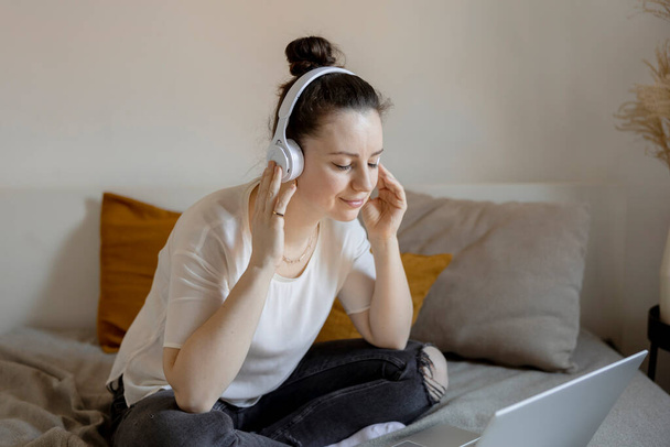 Mladá brunetka sedící doma na posteli s neformálním oblečením a poslouchající hudbu nebo audioknihu. Odpočinek, odpočinek. Čas pro sebe, duševní zdraví. Dívka sní. - Fotografie, Obrázek