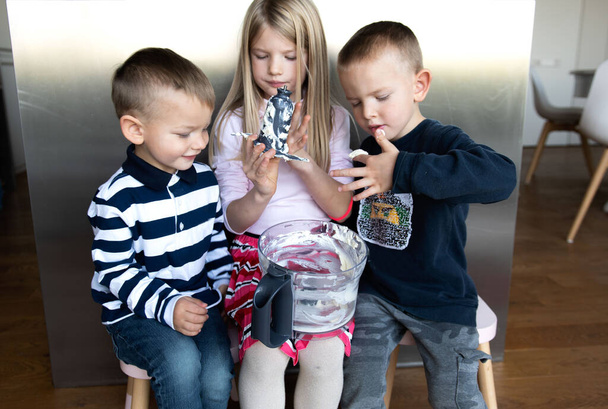 Três crianças na cozinha comem creme doce enquanto a mãe prepara um bolo de férias - Foto, Imagem